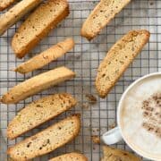 Twelve walnut biscotti assorted on a metal cooling rack with a latte to the side.