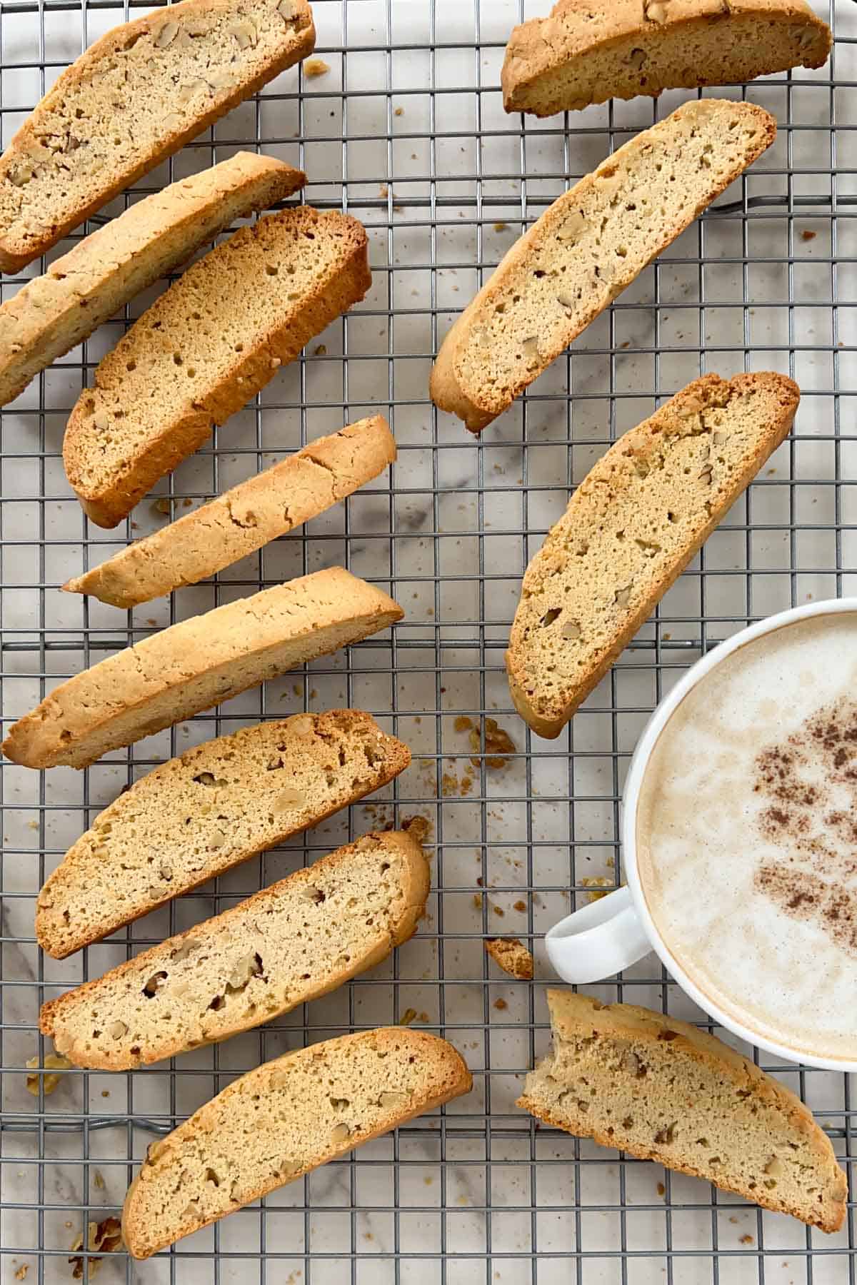 Twelve walnut biscotti assorted on a metal cooling rack with a latte to the side.