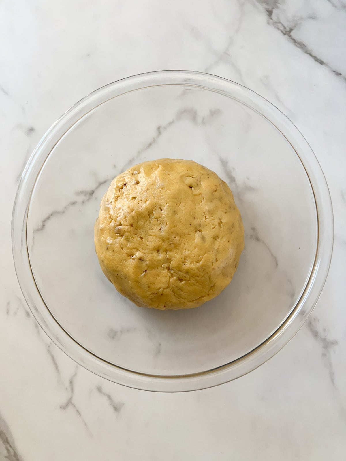 A large glass bowl with a ball of biscotti dough placed on a marble countertop.
