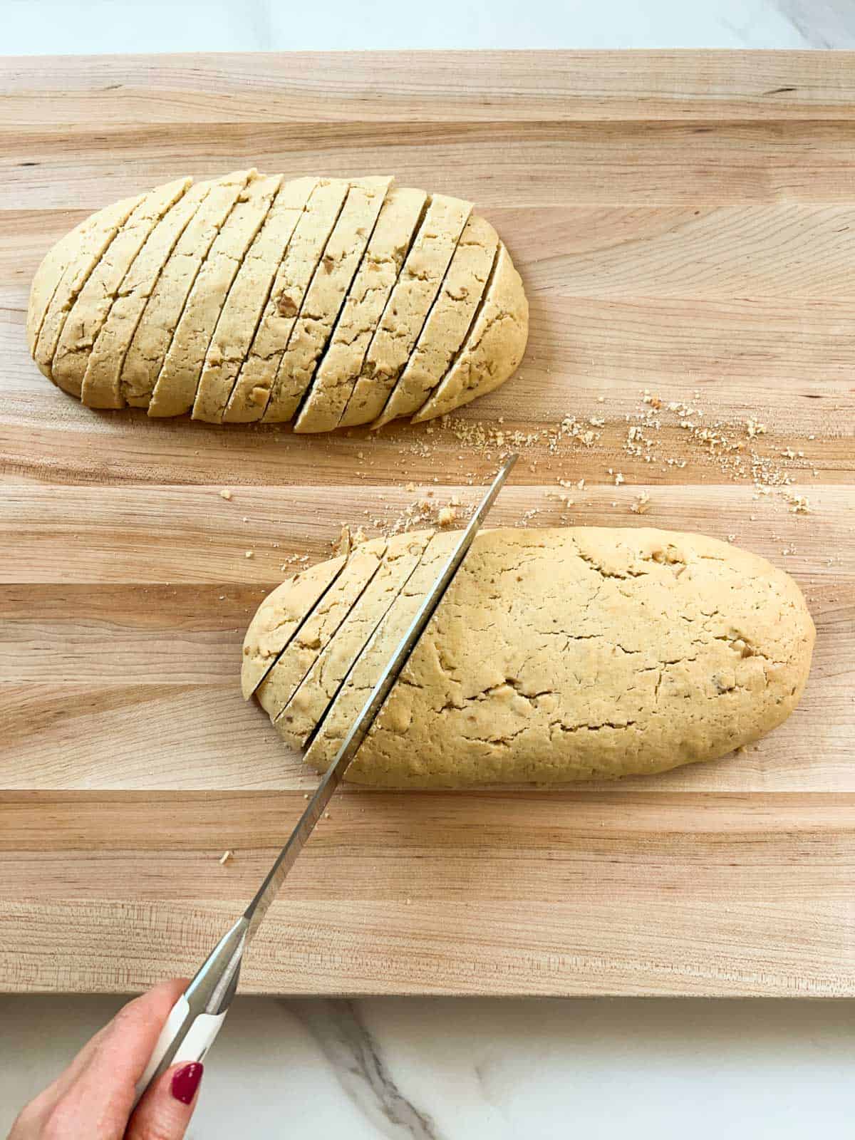 A hand holding a serrated knife is slicing biscotti loaves on a wooden cutting board.